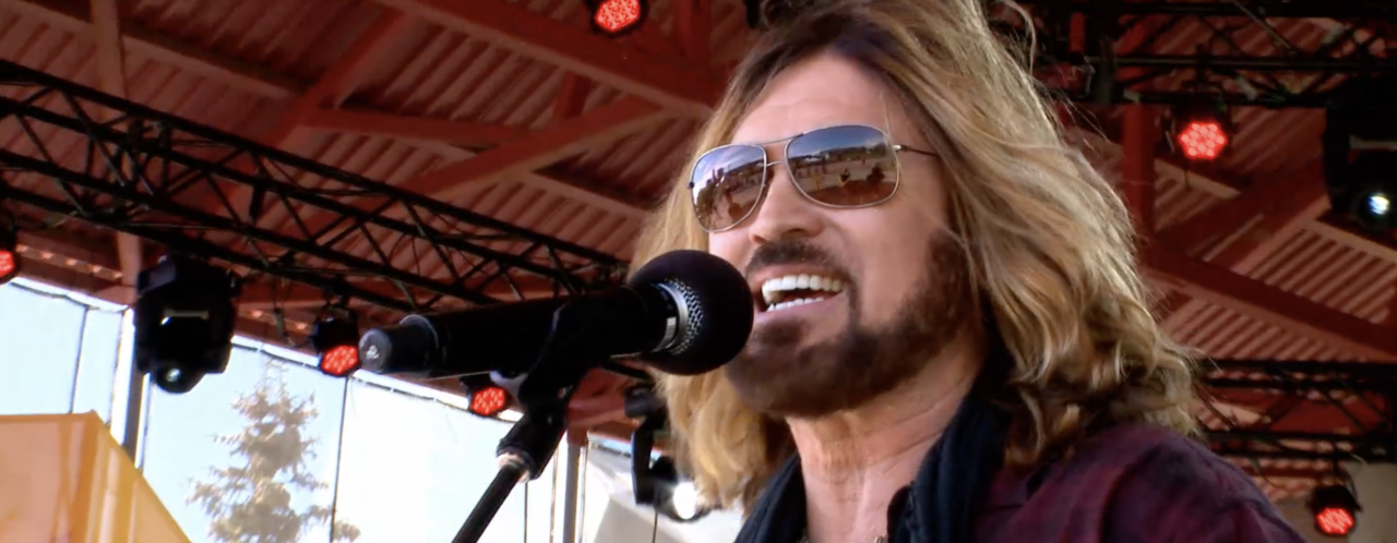 Billy Ray Cyrus preforming on stage on a sunny day outdoors.